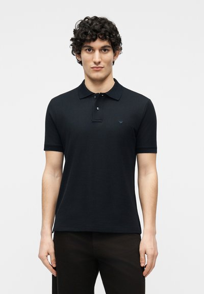Jeune homme aux cheveux bouclés portant un polo noir à manches courtes et un pantalon noir, debout devant un fond blanc uni.