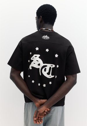 Bărbat cu păr scurt și creț poartă tricou negru cu text alb stilizat și stele pe spate, mâini împreunate la spate, pe fundal simplu.