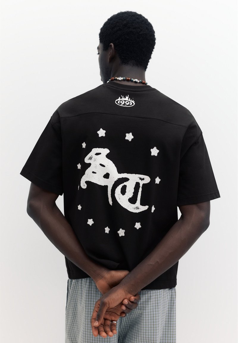 Homme aux cheveux courts et bouclés portant un t-shirt noir avec un texte blanc stylisé et des étoiles dans le dos, mains jointes derrière, sur fond uni.