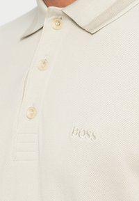 Polo bege claro feito de tecido texturizado com três botões, gola curta e um logótipo em relevo "BOSS" no lado esquerdo.