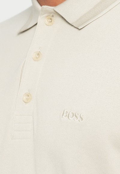Polo beige clair en tissu texturé avec trois boutons, col court et logo "BOSS" en relief sur le côté gauche.