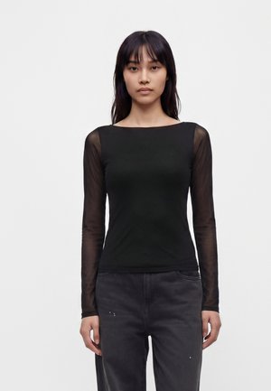 Femme aux longs cheveux foncés portant un haut noir à manches longues transparentes et un pantalon gris foncé sur fond blanc uni.