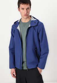 Blå vandtæt jakke med fuld lynlås, justerbar hætte og sorte accenter. Båret over en grøn sweatshirt med elastiske manchetter og sidelommer.