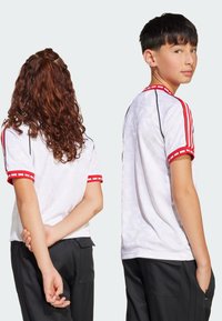 Bijela majica s kratkim rukavima s crvenim i crnim prugastim detaljima, geometrijskim uzorkom i kontrastnim rukavima. Dvoje djece prikazano s leđa.