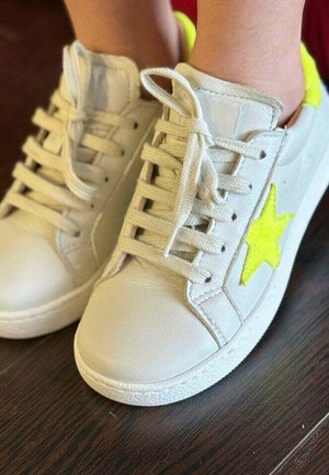 Zapatillas blancas con cordones beige puestas en los pies, con una estrella amarilla brillante en el lateral, sobre un suelo de madera oscura.