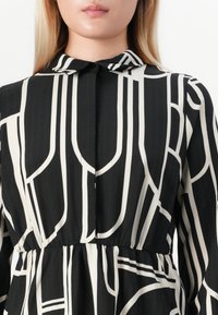 Femme portant une robe noire avec des motifs géométriques verticaux blancs et un petit col, coupée au-dessus de la bouche.