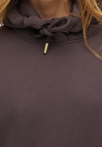 Huvtröja i mörklila tyg med texturerad yta; har en dragsko vid halsen med detaljer i guldton.