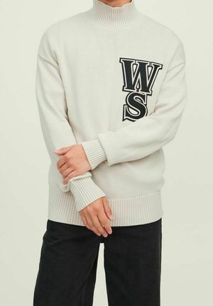 Persona vistiendo un suéter de cuello alto de canalé color crema con letras negras "WS" en el pecho, combinado con pantalones negros, con las manos cruzadas en la cintura.
