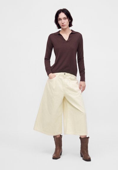 Femme aux cheveux courts et foncés portant un haut marron à manches longues, un large pantalon culotte crème et des bottines marron, debout devant un fond uni.