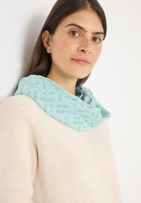 Lichtblauwe, gemusterde infinity-sjaal om de nek gedrapeerd, gemaakt van zachte stof, gecombineerd met een crèmekleurige gebreide trui.