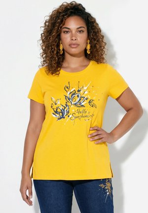 Vrouw met krullend haar draagt een geel "Hello Sunshine" bloemenhemd en blauwe spijkerbroek met bloemenborduursels, poserend met de hand op de heup.