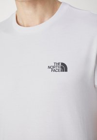 Camiseta de algodón blanca con cuello redondo y un pequeño logo negro de "The North Face" en el lado izquierdo del pecho. Textura suave, diseño sencillo.