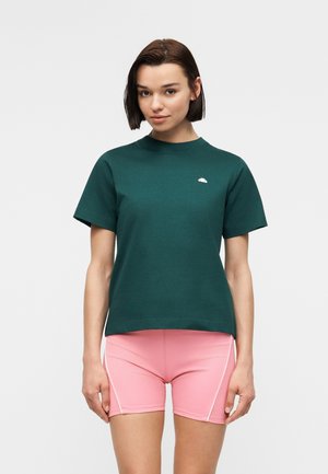 Kvinde med kort mørkt hår iført en mørkegrøn kortærmet top og lyserøde træningsshorts med hvide sidelister, stående mod en ensfarvet baggrund.