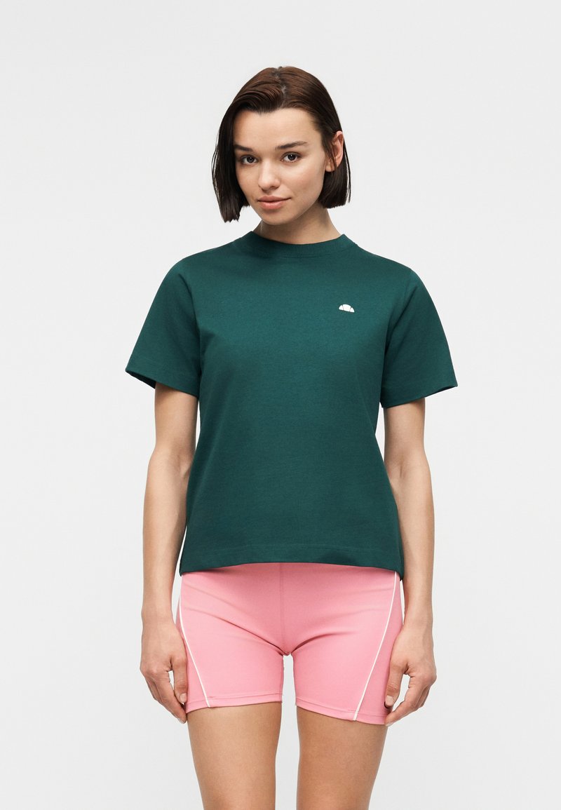 Γυναίκα με κοντά καστανά μαλλιά, φοράει πράσινη κοντομάνικη μπλούζα και ροζ αθλητικά σορτς με λευκές ρίγες στα πλάγια, στέκεται μπροστά σε απλό φόντο.