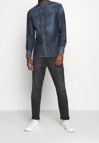 Czarna dżinsowa koszula z guzikami, z dwiema kieszeniami na piersi, jasnymi przeszyciami i kołnierzykiem w kształcie spiczastym, w połączeniu z ciemnymi dżinsami i białymi sneakersami.