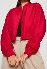 Rode, cropped bomberjack van gladde stof, voorzien van een ritssluiting aan de voorkant, elastische manchetten en zijzakken. Gecombineerd met beige broek.