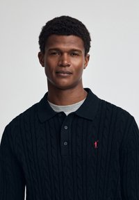 Mörkblå kabelstickad polo med krage, tre knappar och en liten röd logotyp på bröstet. Bärs över en grå rundhalsad skjorta.