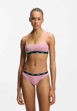 Lichtroze bralette met logoopdruk, verstelbare spaghettibandjes en een zwarte elastische band. Passend lichtroze stringetje met een zwarte tailleband.