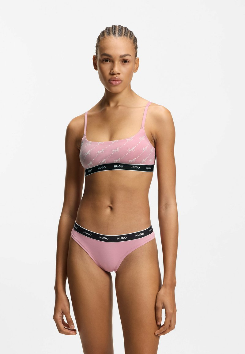 Lyserød bralette med logoprint, justerbare spaghettistropper og en sort elastik. Matchende lyserød thong med sort linning.