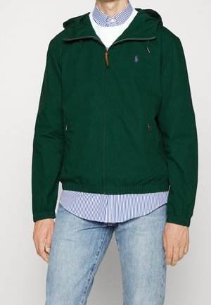 Homme portant une veste zippée verte par-dessus une chemise rayée bleue et blanche et un jean bleu clair, debout devant un fond blanc uni.