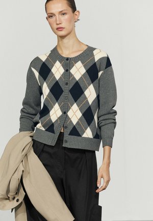 Femme portant un cardigan à motif losanges gris, marine et blanc, tenant un manteau beige dans une main, debout devant un fond clair uni.