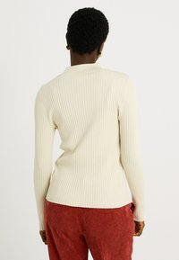 Haut crème côtelé à manches longues avec un col montant, une silhouette ajustée et une légère élasticité, associé à un pantalon en velours côtelé rouge.