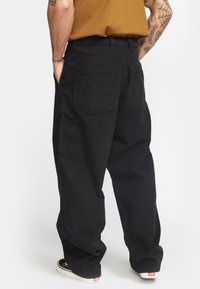 Pantalones cargo negros con un ajuste holgado, que cuentan con múltiples bolsillos, una cinturilla plana y una tela suave y texturizada. Se aprecian detalles de zapatillas blancas.