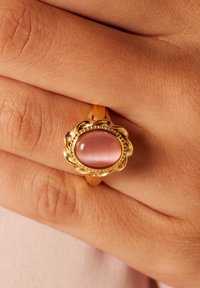 Gouden ring met een roze cabochonsteen in een bloemvormige setting, met een gladde band en een gestructureerd oppervlak op een hand.