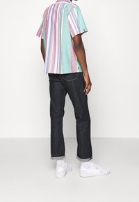 Uomo in camicia a righe multicolori con maniche corte, jeans scuri con risvolto, calze bianche e sneakers bianche, in piedi contro un muro bianco.