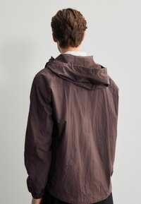 Jachetă ușoară cu glugă, de culoare mauve închis, fabricată dintr-un material texturat. Prezintă mansete elastice și o croială lejeră, cu detalii vizibile ale cusăturii.