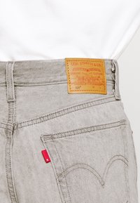 Los jeans de mezclilla gris presentan una etiqueta de cuero en la cintura, con un acento de logo rojo y detalles bordados en los bolsillos traseros.