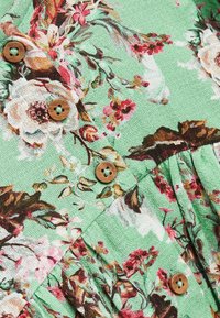 Textilie s květinovým vzorem v zelené barvě s texturovaným povrchem, zdobená vícero barevnými květinami a dvěma dřevěnými knoflíky se starými otvory ve tvaru hvězdy.