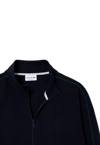 Dunkelblaues Zip-Jackett mit strukturiertem Obermaterial, grünen Akzenten an den Schultern und einem gerippten Kragen. Enthält ein Lacoste-Logo-Etikett innen.