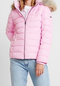 Rosa Pufferjacke mit gefütterter Kapuze, gestepptem Design, Frontreißverschluss und seitlichen Taschen. Kombiniert mit hellblauen Jeans.