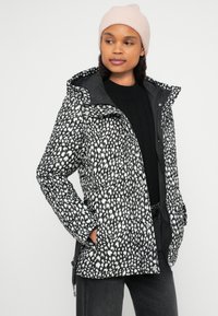 Μαύρο και άσπρο μπουφάν-φουσκωτό με μοτίβο, κουκούλα, φερμουάρ και κλείσιμο με κουμπιά, φορεμένο πάνω από ένα μαύρο πουλόβερ. Το μοντέλο φοράει ροζ σκουφάκι.