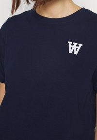 Tmavě modré bavlněné tričko s kulatým výstřihem, krátkým rukávem a bílým logem s písmeny "W" na hrudi.