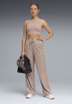 Kobieta w beżowym topie Puma i pasiastych spodniach, trzymająca kwiecistą czapkę i czarny ręcznik, stojąca na szarym tle.