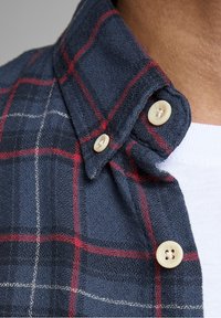 Námořnická flanelová košile s červeným a bílým kostkovaným vzorem, límcem na knoflíky a světle zbarvenými knoflíky. Viditelný detail texturované látky.