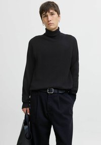 Jersey de cuello alto negro acanalado con mangas raglán. Combinado con pantalones negros y un cinturón a juego. Se sostiene un bolso negro elegante con una mano.