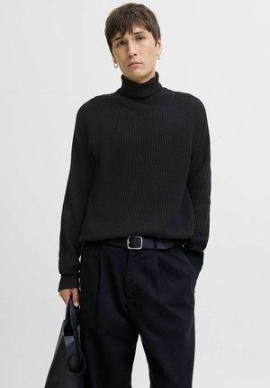 Jersey de cuello alto negro acanalado con mangas raglán. Combinado con pantalones negros y un cinturón a juego. Se sostiene un bolso negro elegante con una mano.