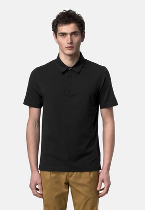 Giovane uomo con polo nera a maniche corte e pantaloni color sabbia, in piedi davanti a uno sfondo grigio chiaro uniforme.