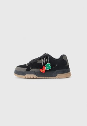 Svart og grå sneaker med en beige gummisåle, tykke snørebånd og to dekorative anheng: et rødt hjerte merket "Demon" og et grønt dollartegn.