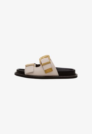 Sandalias deslizantes de cuero crema con dos hebillas doradas, suela negra y detalle de costura blanca a lo largo del borde. Logotipo de la marca mostrado de manera prominente.