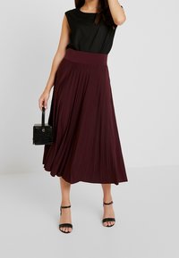Gonna plissettata bordeaux con una cintura ampia, abbinata a una maglietta nera. Porta una piccola borsa nera testurizzata e indossa sandali neri con tacco.