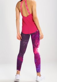 Donna in piedi con un canottiera Nike rosa brillante senza schienale e leggings a fantasia galattica viola e rosa, abbinati a sneakers bianche.