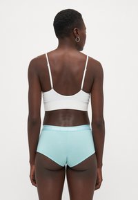 Bela rebrasta bralette in svetlo modri bokserici z širokim pasom, ki nosi logo "PUMA", prikazana s hrbtne strani na enobarvni podlagi.