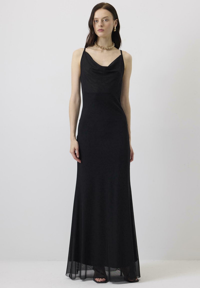 Γυναίκα που φοράει μακρύ μαύρο φόρεμα χωρίς μανίκια με λεπτές τιράντες, χρυσό κολιέ, σκουλαρίκια κρίκους και μαύρα σανδάλια ανοιχτά στα δάχτυλα, στέκεται μπροστά σε απλό φόντο.