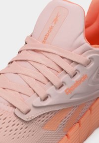 Κοντινό πλάνο ροζ αθλητικού παπουτσιού Reebok με υφασμάτινο άνω μέρος με υφή πλέξης, κορδόνια στο ίδιο χρώμα και επενδεδυμένη γλώσσα με λογότυπο, το οποίο στέκεται σε ροδακινί σόλα.