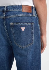 Vaqueros de denim en azul oscuro con un parche de cuero en la cinturilla y un logo triangular rojo en el bolsillo trasero. Tejido texturizado.