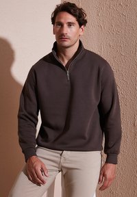 Bruine rits-trui met lange mouwen, zachte textuur, ribbelboorden en halslijn, gecombineerd met lichtbeige broeken.